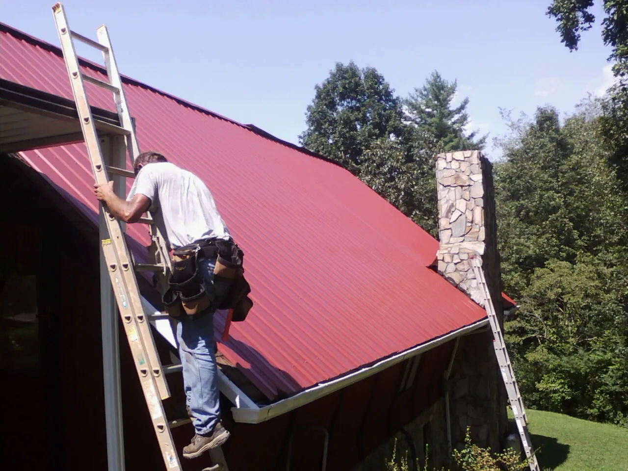 Roofer on a ladder beside a red metal roof and stone chimney — Soffit & Fascia Repair by Roofing Brothers in Norwood